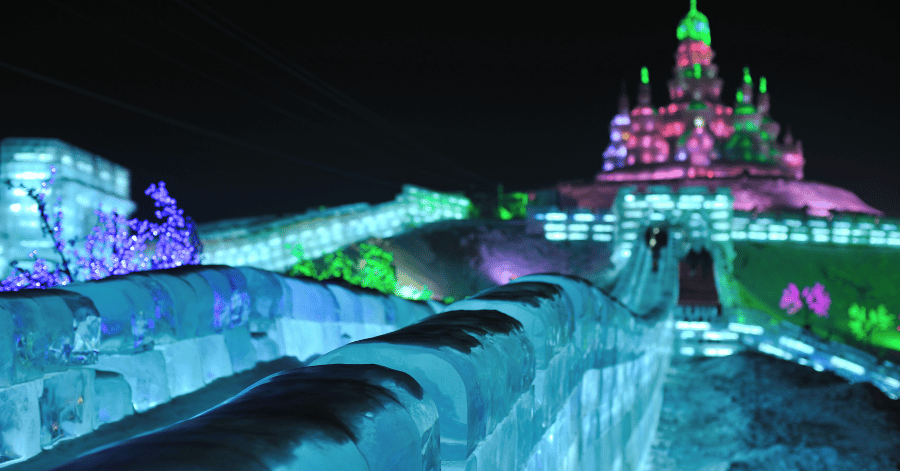 A long illuminated ice slide leading toward a floodlit pink-and-green ice castle at night