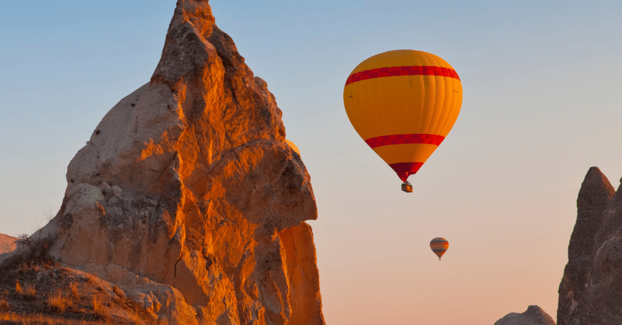 A yellow hot air balloon floating past a tall Cappadocia rock pinnacle at dusk