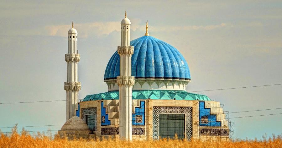 A mosque with a blue tiled dome and two white minarets above dry golden grassland