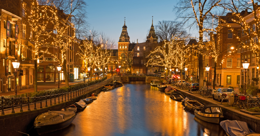 An Amsterdam canal at dusk, trees strung with fairy lights and boats moored along both banks