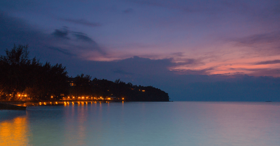 Pink and blue dusk sky over calm water with lights along a dark treelined shore