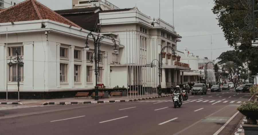 Motorcyclist passing white colonial buildings along a wide street in central Bandung