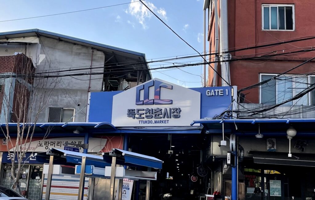 Blue and white Ttukdo Market gate sign above the entrance to a covered market alley