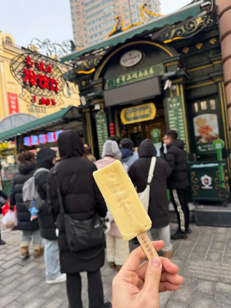 A hand holding a pale yellow Madie'er ice cream bar outside the green Modern ice cream shop, people queuing in winter coats