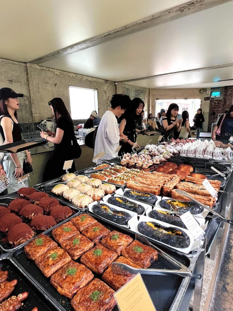 Trays of pastries and pull-apart breads on a counter with customers queuing behind