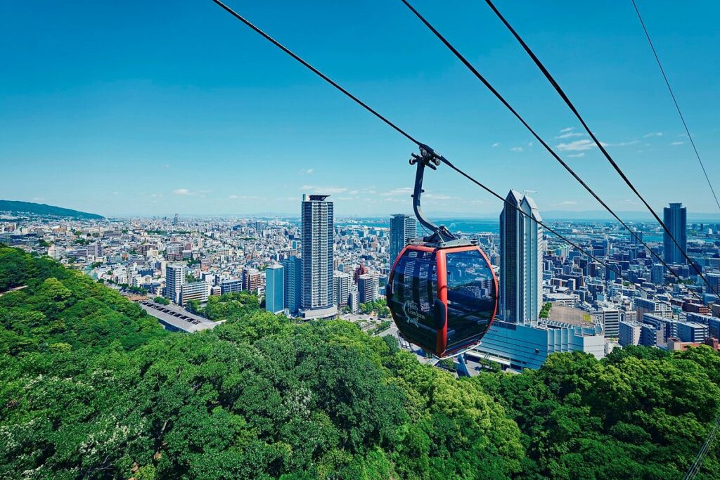 Red ropeway gondola climbing above Kobe with the city skyline and bay behind