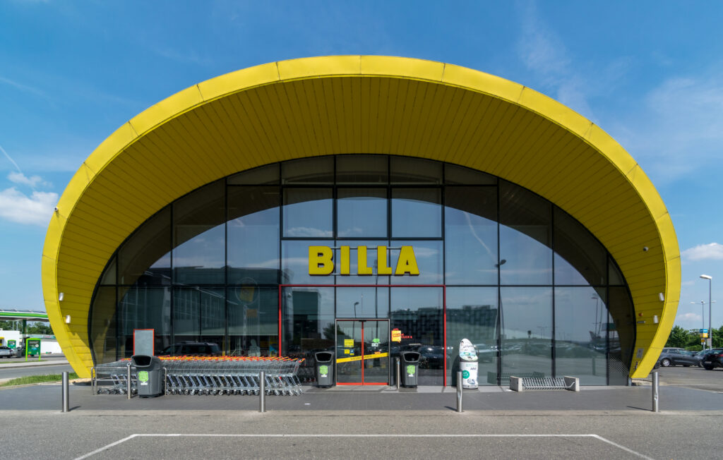 The curved yellow facade of a BILLA supermarket with trolleys outside the entrance