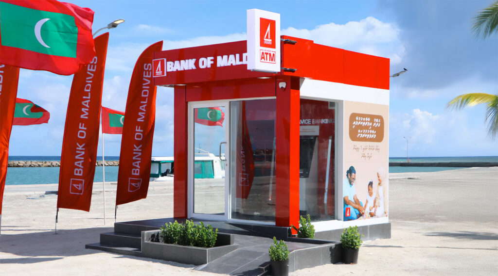 A standalone Bank of Maldives ATM kiosk on a beachfront with Maldives flags
