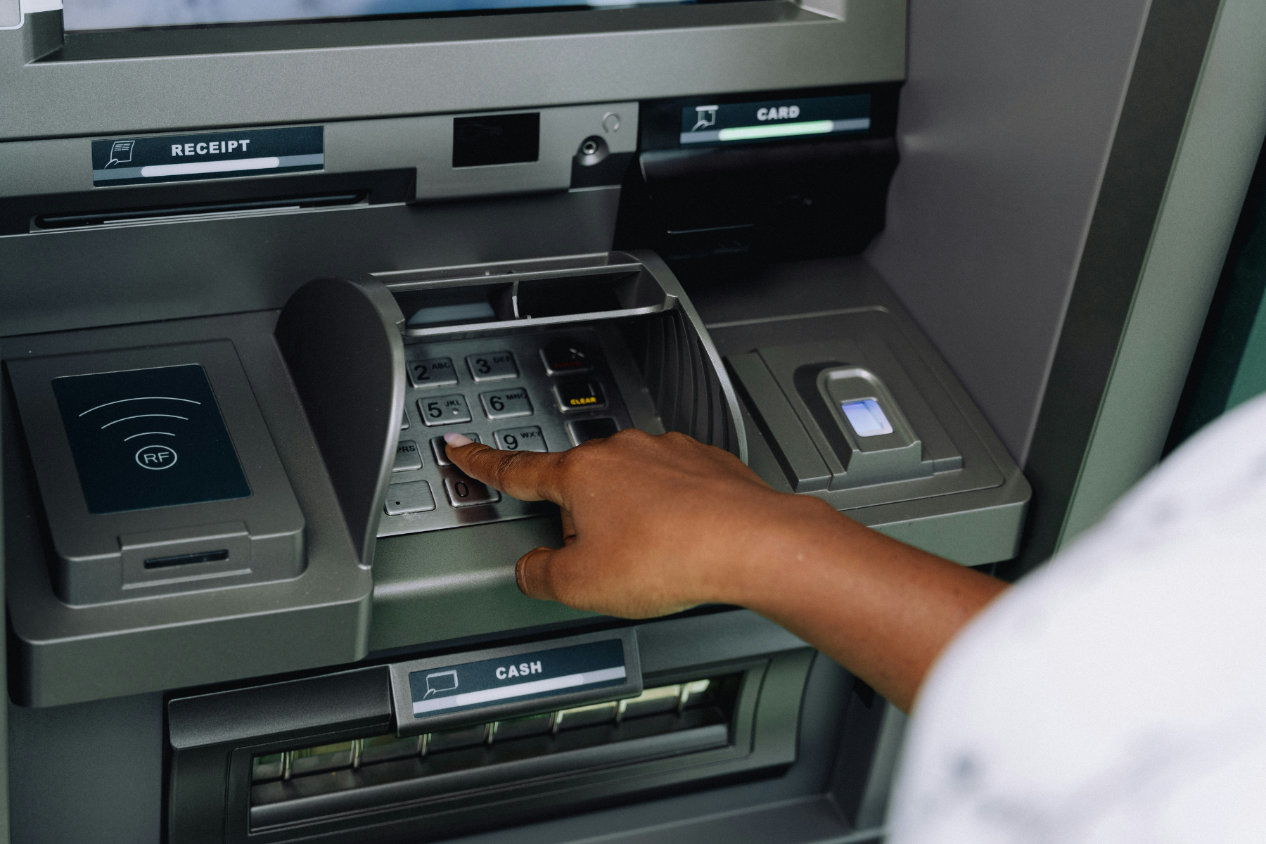 A hand entering a PIN on an ATM keypad beside the card, receipt and cash slots