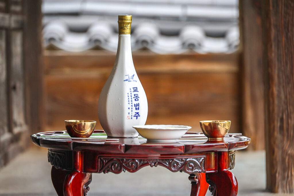 White ceramic bottle of Gyodong Beopju rice wine with two brass cups on a red lacquer table