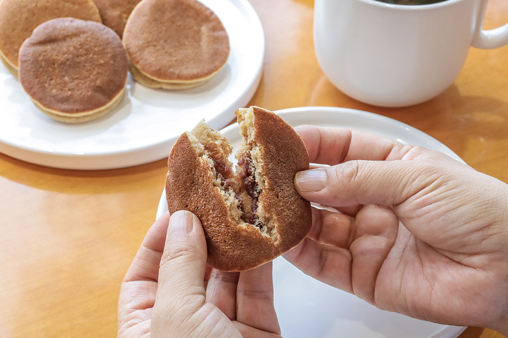 Hands splitting a barley bun to show its red bean filling, with more on a plate and coffee