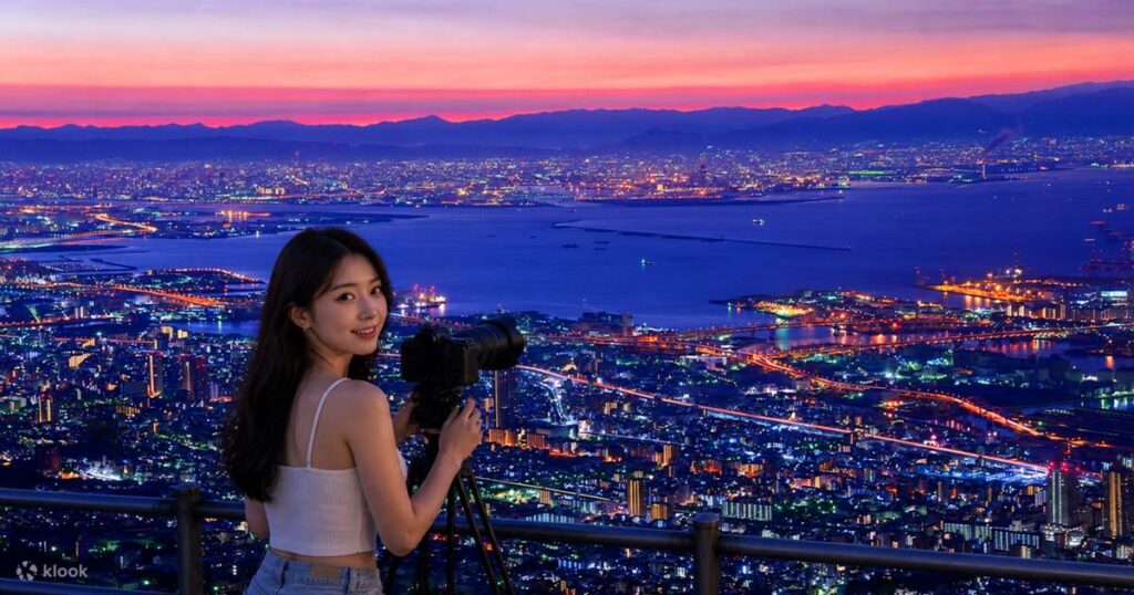 A visitor photographing the sunset skyline from a mountain viewpoint above Kobe