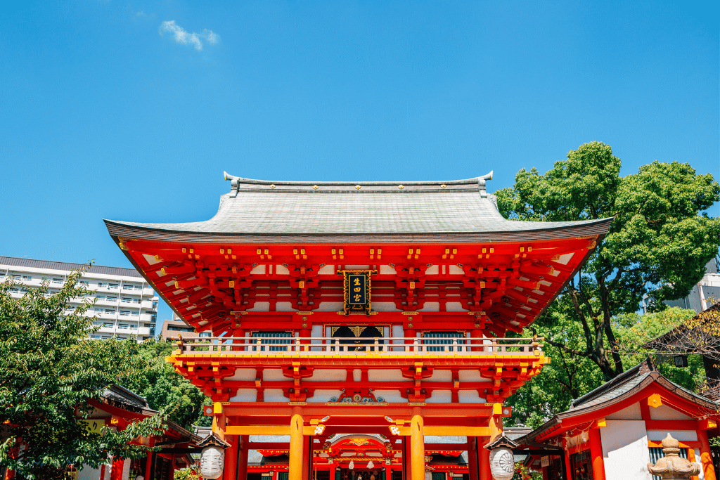 The red gate of Ikuta Shrine framed by trees against a blue sky