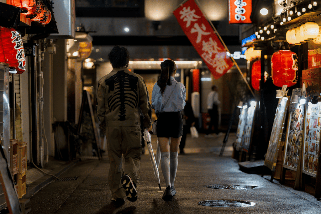 Two people walking past lantern-lit izakaya signage in a Kobe backstreet at night