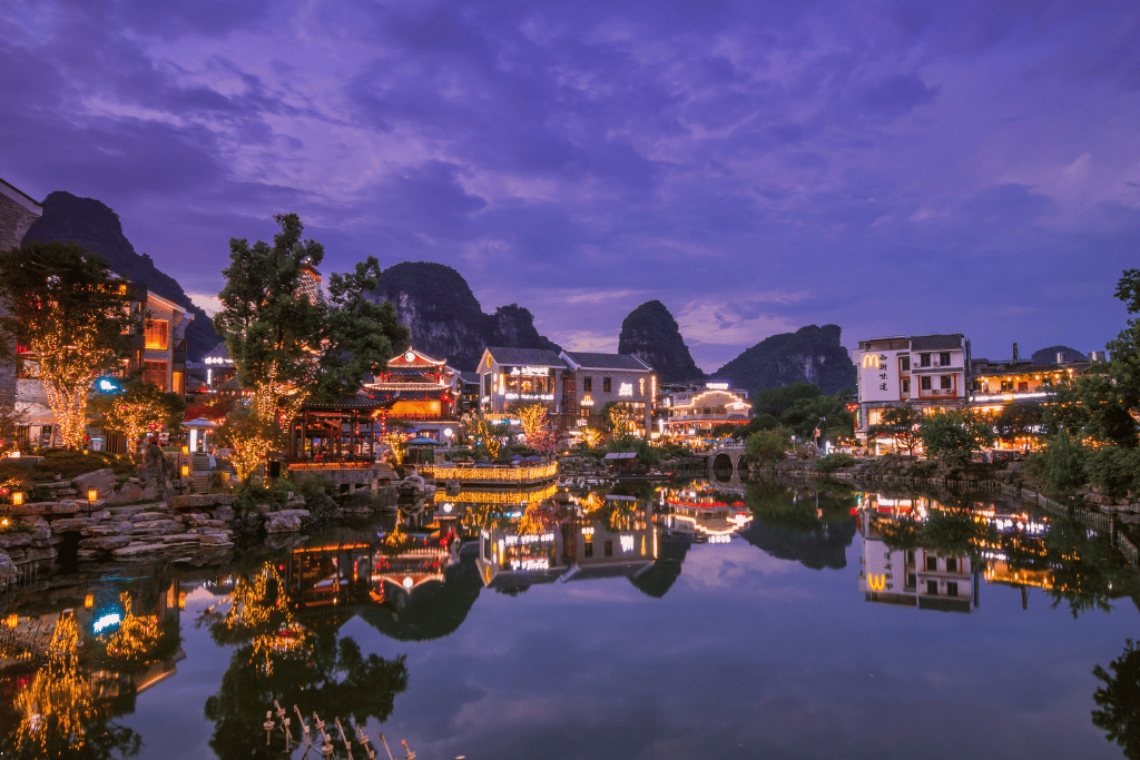 Yangshuo's riverside restaurants and shops lit up at dusk, karst peaks silhouetted behind, reflected in the water
