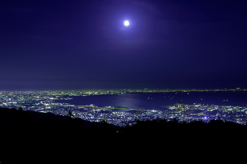 A full moon rising over Kobe Bay's night skyline seen from a hillside