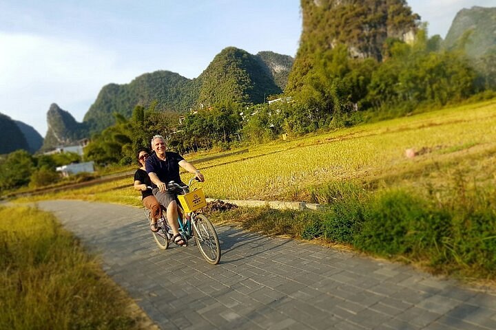 Two cyclists riding a shared bicycle along a paved path through rice fields on the Ten-Mile Gallery