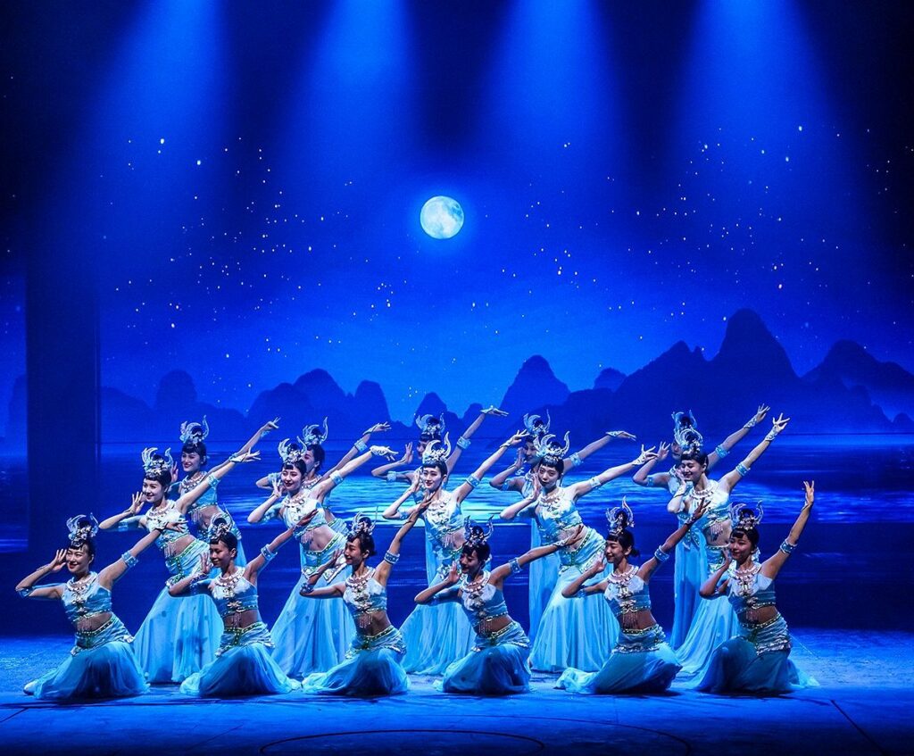 Dancers in blue costume performing on stage at Guilin Romance Park, a painted karst-mountain backdrop behind