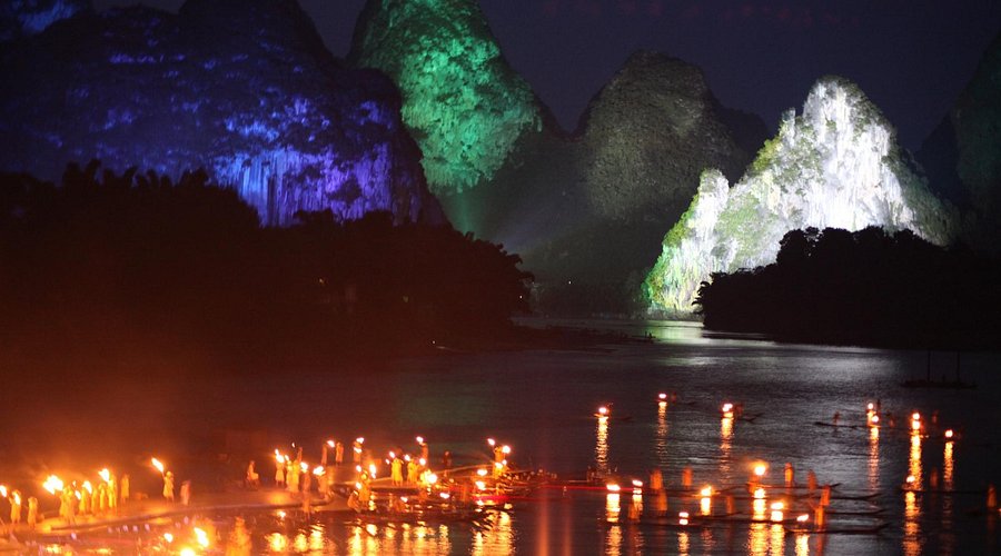 Torch-lit rafts on the Li River at night with karst peaks lit blue and green as a backdrop