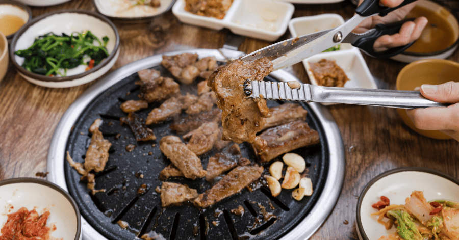 Tongs lifting grilled pork off a charcoal Korean BBQ grill in Johor Bahru