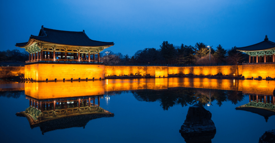 Lit pavilions and stone wall of Wolji Pond mirrored in still water at blue hour