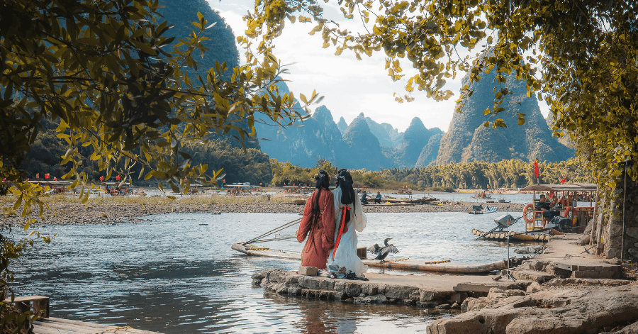 Two women in traditional red and white dress standing by the Li River with bamboo rafts