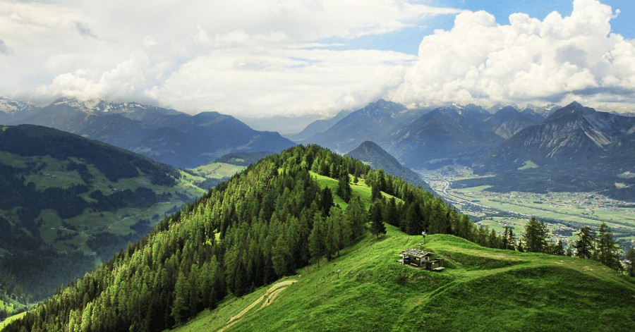 A forested ridge and green pasture above an Austrian alpine valley, mountains beyond