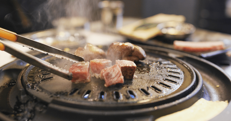 Tongs grilling marinated pork cubes on a Korean BBQ grill plate