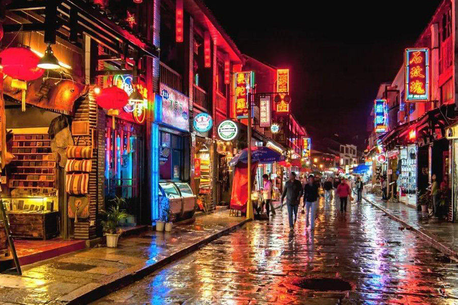 A pedestrian street at night lined with red lanterns and neon shop signs, pavement wet with rain