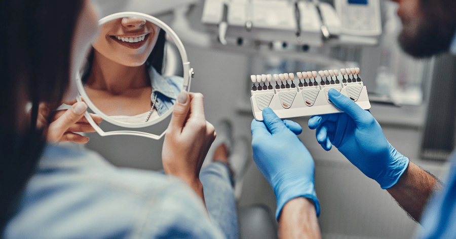 Dentist in blue gloves holding a tooth shade guide while a patient checks her smile in a hand mirror
