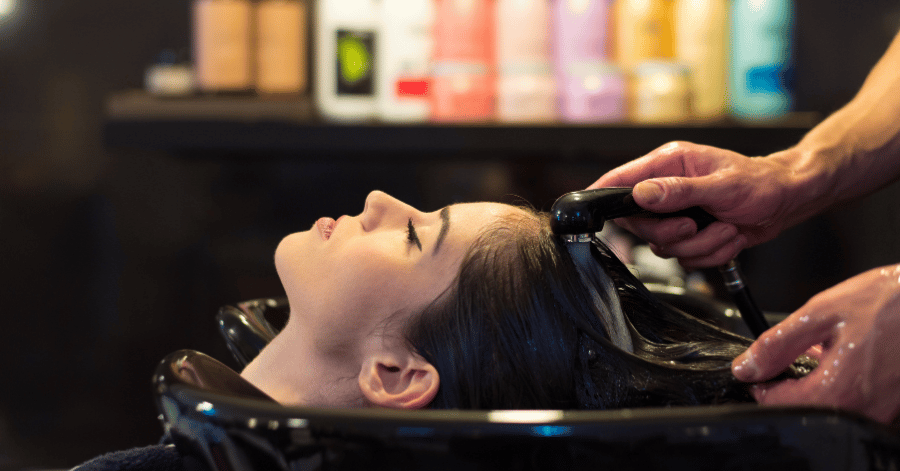 Close-up of a woman having her hair washed at a salon basin, with hair products in the background
