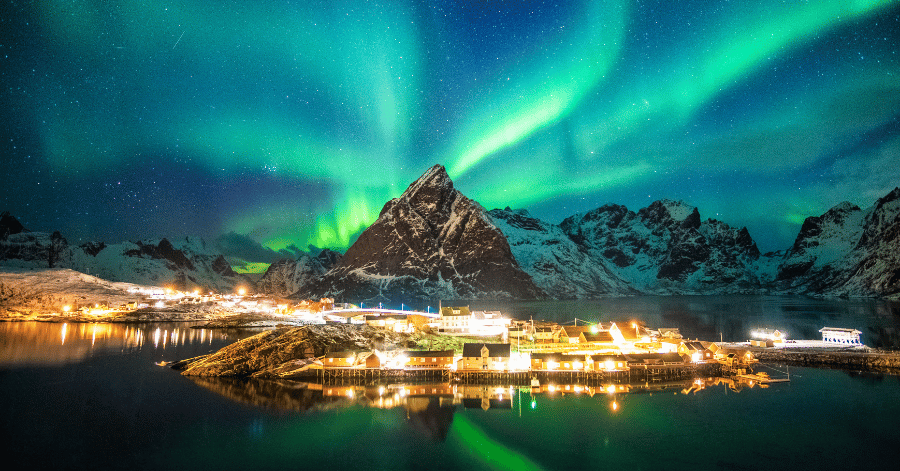 Northern lights above a snow-capped peak and a lit fishing village reflected in still water