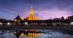 Pha That Luang's golden stupa illuminated at dusk in Vientiane, reflected in a puddle on the plaza below