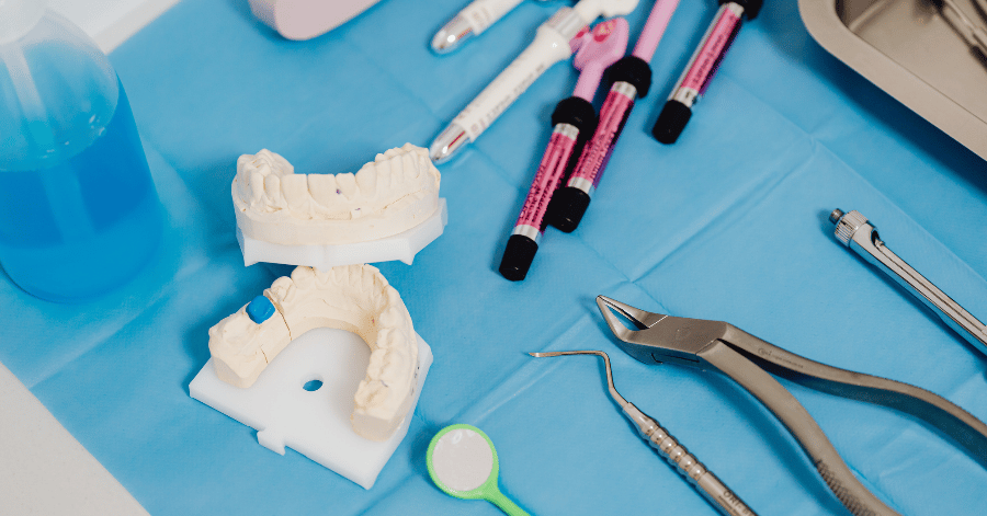 Dental mirror, extraction forceps, filling syringes and two tooth models laid out on a blue surface