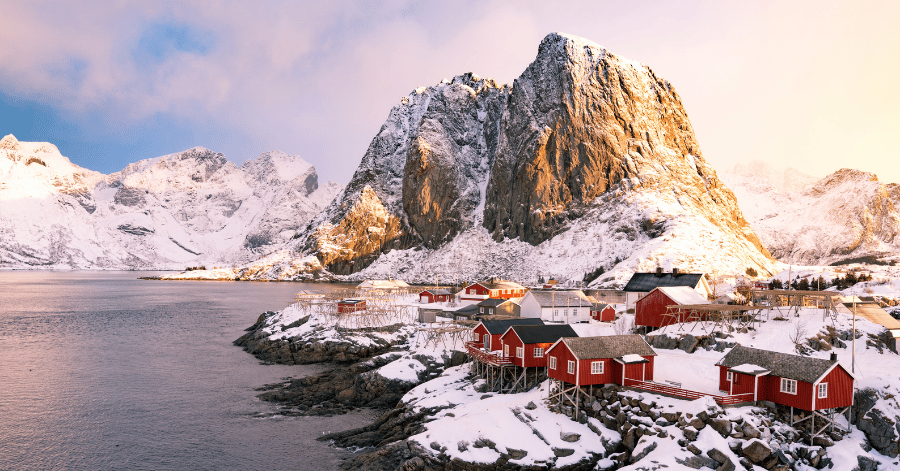 Red wooden cabins on a snowy shore below a sunlit mountain peak