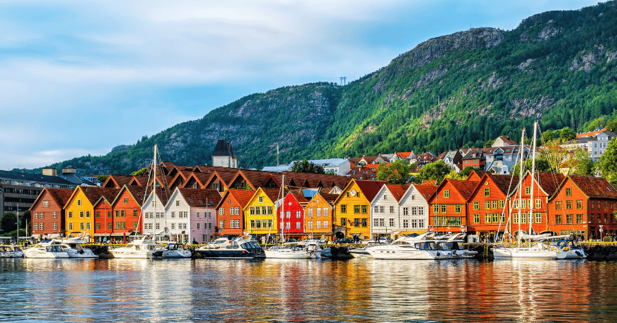 Row of colourful wooden waterfront buildings with boats moored in front and a forested hill behind