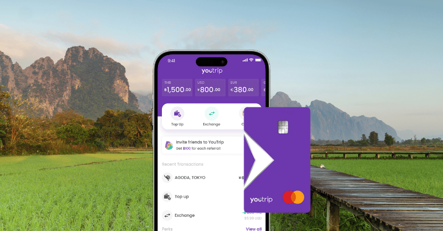 A phone showing the YouTrip app's currency balances next to a YouTrip card, set against Vang Vieng's rice fields and karst hills